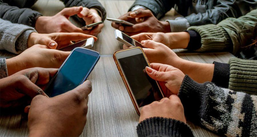 دست چند نفر که کنارهم دور میز نشسته‌اند و موبایل در دست دارند در تصویر مشخص است. 