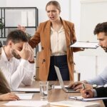 دو خانم و دو آقا با ناراحتی دور میز کار نشسته‌اند و یک خانم ایستاده و در حال فریاد زدن سر آن‌ها است.