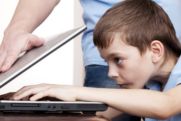 پسربچه‌ای جلوی لپتاپ نشسته و دستش روی کیبورد است و مردی که در کنار او ایستاده در حال بستن لپتاپ است.